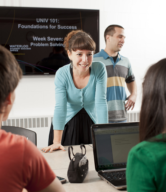 Success coach Andrea Prier engages with a pair of students as she stands in front of a presentation screen featuring UNIV 101 slides.